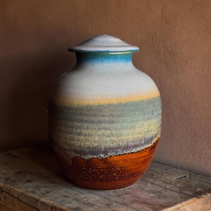 May include: A ceramic urn with a lid, featuring a gradient of colours. The top is light blue, transitioning through yellow, green, and grey to a brown base. The urn is displayed on a wooden surface. The height is approximately 30 cm.