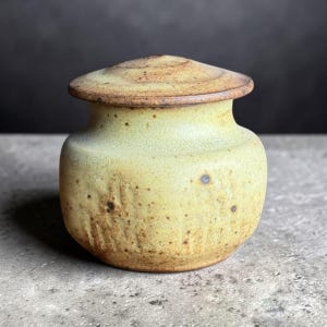 Könnte beinhalten: Ein kleiner, handgefertigter Keramiktopf mit Deckel. Der Topf ist hellgelb mit braunen Sprenkeln und einem dunkleren braunen Rand und Deckel. Der Topf hat einen abgerundeten Körper und eine leicht strukturierte Oberfläche.