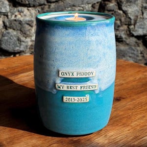 May include: A teal blue ceramic urn with a white glaze and a small teal blue lid. The urn has three white rectangular labels with the text "ONYX PIDDLY", "MY BEST FRIEND", and "2013-2025". A small white tea light candle is burning in the lid.