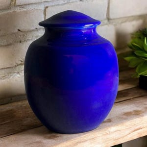 Puede incluir: Una urna de cerámica azul oscuro con una forma redonda y lisa y una tapa. La urna es un diseño clásico y sería una hermosa adición a cualquier hogar.