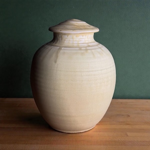 Op de afbeelding: Een crèmekleurige keramische urn met een deksel. De urn heeft een afgeronde body en een licht gestructureerd oppervlak. Het deksel heeft een bijpassende crèmekleur en zit bovenop de urn. De achtergrond is donkergroen.