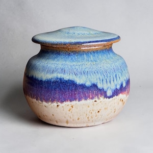 May include: A round ceramic jar with a matching lid, featuring a reactive glaze that transitions from a light blue top to a deep purple middle section, ending in a speckled cream-colored base with a smooth, glossy finish.