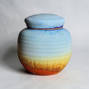 Peut inclure: Un pot en céramique avec couvercle présentant un émail dégradé passant d'un bleu clair sur le dessus à un jaune au milieu et une base orange foncé. Le récipient a une forme arrondie et bombée avec des stries horizontales sur le corps.