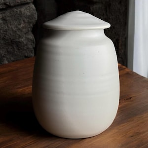 Puede incluir: Un tarro de cerámica blanco con tapa. El tarro tiene una forma redonda y lisa y una superficie ligeramente texturizada.