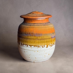 May include: A ceramic jar with a lid, featuring a multi-toned glaze that transitions from a deep orange at the top to a golden yellow in the middle, and a speckled white base. The glaze exhibits a distinct drip pattern where the colors meet.