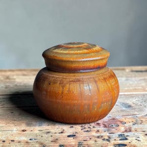Peut inclure: Un pot en céramique rond avec un couvercle assorti, présentant une glaçure brillante dans des tons orange brûlé, brun profond et des touches de jaune doré. La surface présente de légères stries verticales et une silhouette arrondie.