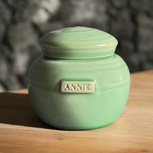 May include: A light green ceramic jar with a lid and the name "ANNIE" written on the front.