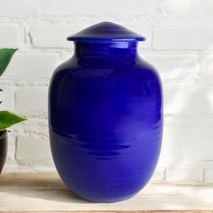 Puede incluir: Una urna de cerámica azul oscuro con tapa. La urna tiene un acabado liso y brillante.