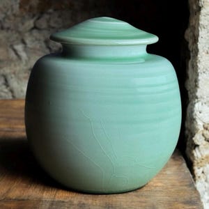 Puede incluir: Una urna de cerámica verde claro con una forma redonda y lisa y una tapa. La urna tiene un acabado texturizado sutil.