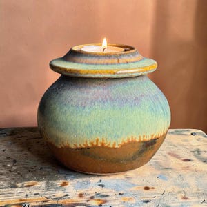 May include: A round ceramic candle holder with a wide, flat rim and a tapered body. The glaze features a gradient of earthy brown at the base, transitioning into a soft sage green and a hint of purple near the top. A lit tea light candle sits in the center.