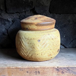 Könnte beinhalten: Eine weiße Keramiktasse mit schwarzem Henkel und dem Text "But First, Coffee" in fetter, schwarzer Schrift. Die Tasse steht vor einem verschwommenen Hintergrund, der eine gemütliche Umgebung suggeriert.