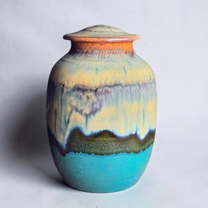 Op de afbeelding: Een keramische urn met deksel, voorzien van een meerkleurig glazuur dat verloopt van een feloranje rand naar een lichtgeel middengedeelte, gevolgd door een donker olijfgroene band en een effen, levendige turquoise basis met gladde afwerking.