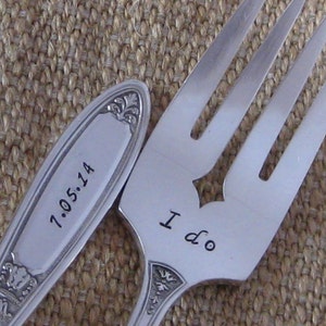 May include: Close-up of two silver utensils, a fork and a spoon, with engraved text. The fork reads "I do" and the spoon reads "7.05.14". Both utensils have decorative floral patterns.