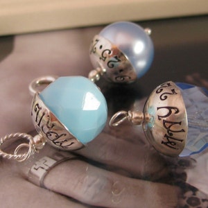 Puede incluir: Tres colgantes de plata con cuentas de cristal azul. Cada colgante tiene una banda de plata con las palabras "July 26" grabadas.