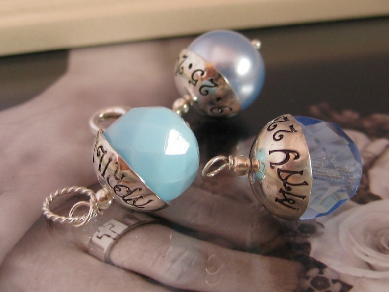 Puede incluir: Tres colgantes de plata con cuentas de cristal azul. Cada colgante tiene una banda de plata con las palabras "July 26" grabadas.