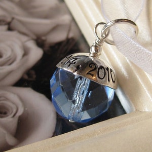 Puede incluir: Un charm de plata con un cristal azul y la fecha "12.9.2010" grabada en &eacute;l.