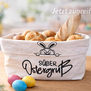Könnte beinhalten: Ein Stoffkorb gefüllt mit Brötchen und einem Baguette. Der Korb hat eine Hasen-Grafik und die Worte "Süßer Ostergruß". Bunte Ostereier liegen vor dem Korb.