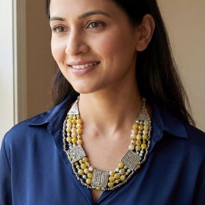 Puede incluir: Collar de múltiples hilos con cuentas amarillas y blancas, acentuado con cuadrados y triángulos de metal plateado. El collar se lleva sobre una camisa azul oscuro.
