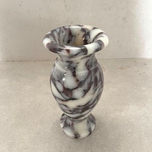 May include: A small, polished stone vase featuring a marbled pattern with deep purple and white veins. The vase has a flared rim, a narrow neck, a rounded body, and a circular base, showcasing a smooth, glossy finish throughout the entire piece.
