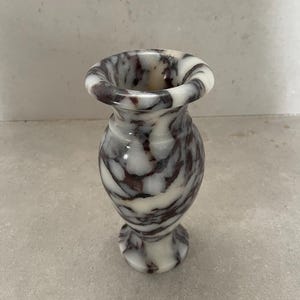 May include: A small, polished stone vase featuring a distinct marbled pattern with swirling veins of deep purple and dark brown set against a creamy white background. The vase has a flared rim, a narrow neck, and a rounded body on a pedestal base.