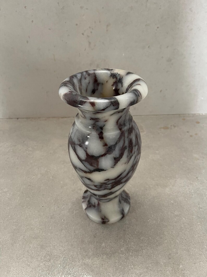 May include: A small, polished stone vase featuring a distinct marbled pattern with swirling veins of deep purple and dark brown set against a creamy white background. The vase has a flared rim, a narrow neck, and a rounded body on a pedestal base.