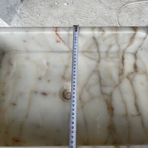 This luxurious Calacatta Gold marble sink features a standard drain hole with a back panel and a striking, elegant design with prominent grey and yellow veins on the front. It can be wall-mounted or countertop-mounted. The front has a fluted design. Dimensions: 24 inches long, 5 inches high, 14 inches deep, panel height 8 inches.