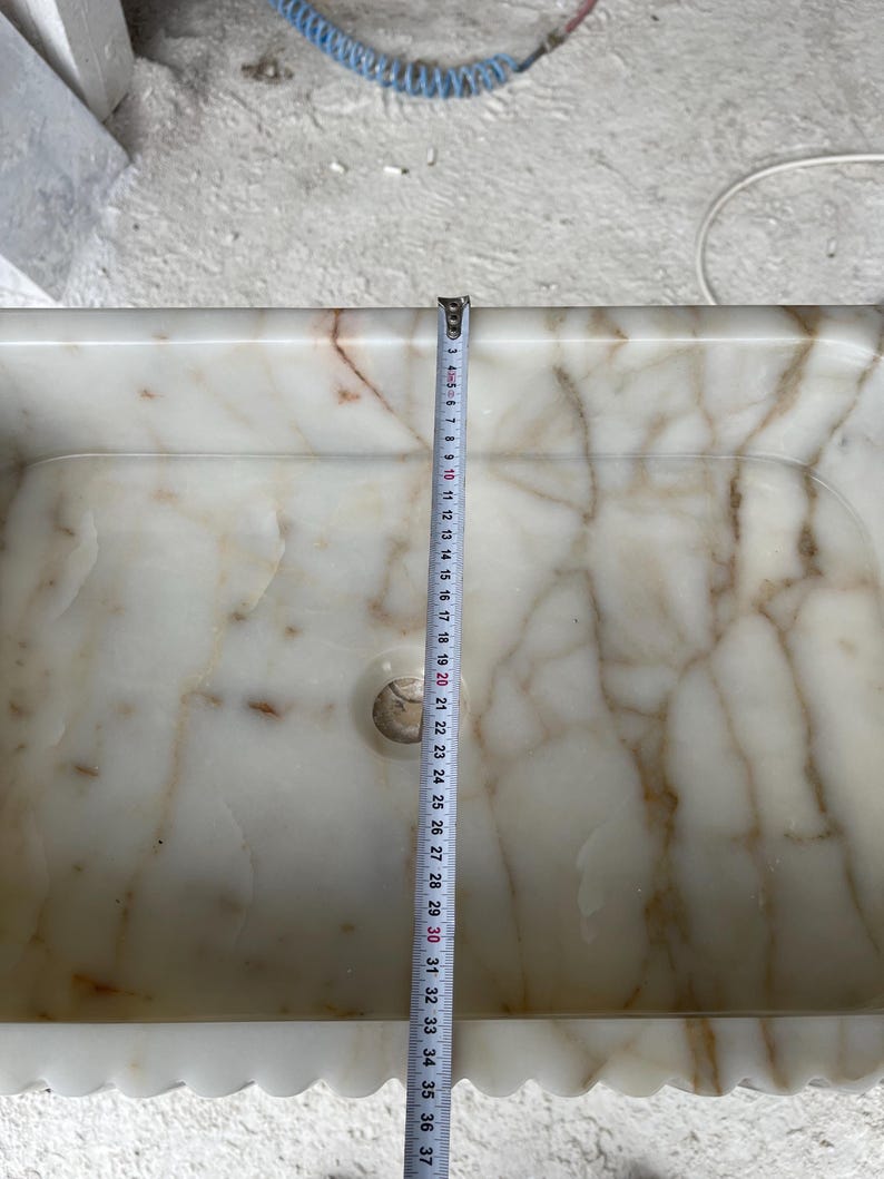 This luxurious Calacatta Gold marble sink features a standard drain hole with a back panel and a striking, elegant design with prominent grey and yellow veins on the front. It can be wall-mounted or countertop-mounted. The front has a fluted design. Dimensions: 24 inches long, 5 inches high, 14 inches deep, panel height 8 inches.