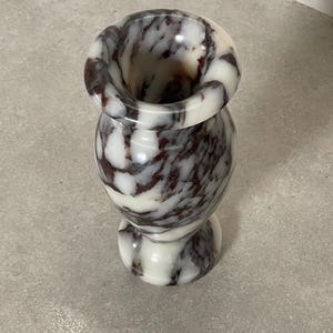 May include: A small, polished stone vase featuring a rounded body and a flared rim. The surface displays a marbled pattern with swirling veins of deep burgundy and dark brown set against a smooth, creamy white background throughout the piece.