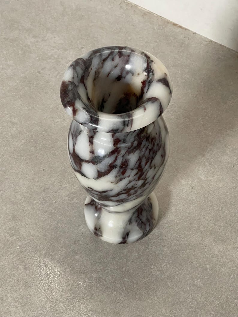 May include: A small, polished stone vase featuring a rounded body and a flared rim. The surface displays a marbled pattern with swirling veins of deep burgundy and dark brown set against a smooth, creamy white background throughout the piece.