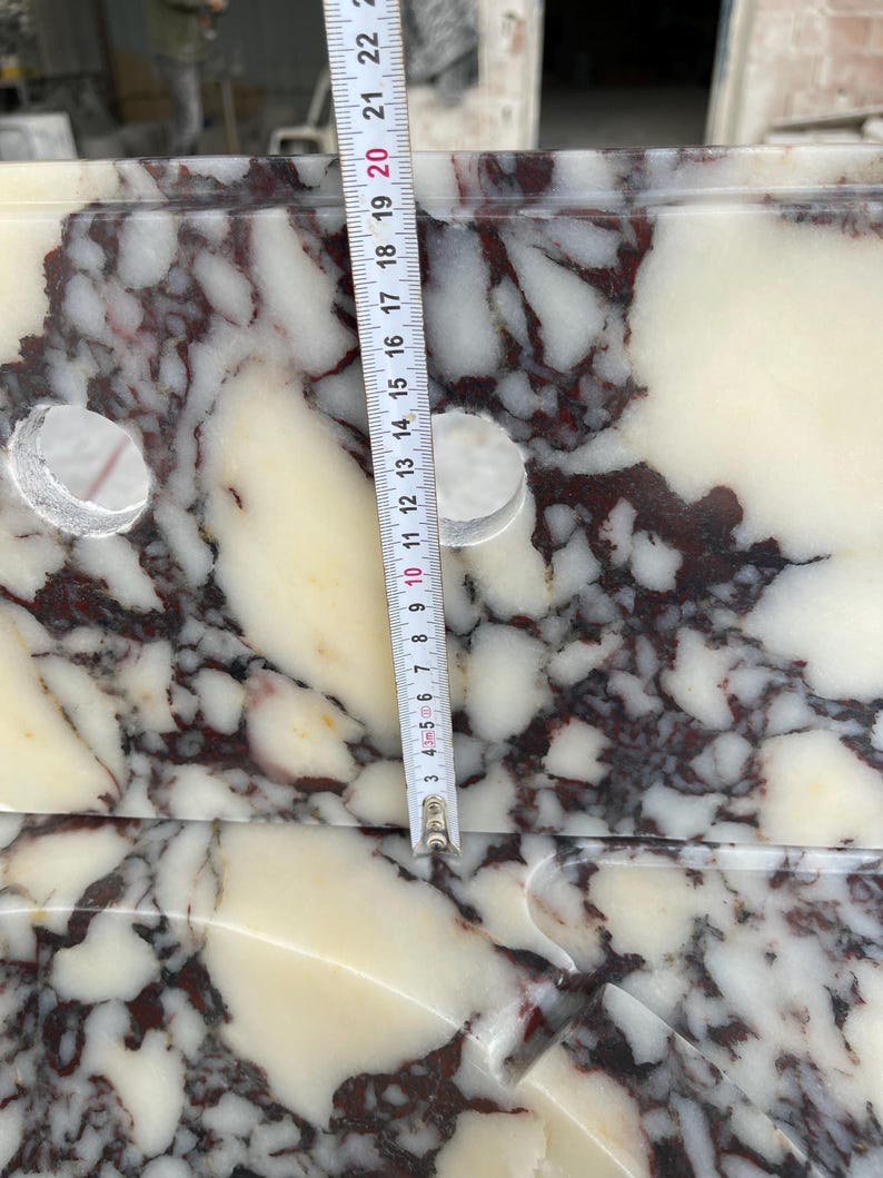 Op de afbeelding: Een gepolijste marmeren wastafel met een witte basis en donkerrode en bruine aders. Een verticaal meetlint ligt op het oppervlak en geeft een lengte van ongeveer 22,2 cm aan tussen de twee ronde gaten voor de kraan.