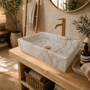 May include: A rectangular white marble vessel sink with brown veining, featuring a brushed gold faucet and a matching gold drain stopper, set upon a light-toned wooden vanity surface in a bathroom setting with neutral-colored decor elements.