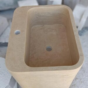 May include: A rectangular beige stone pedestal sink with rounded corners, featuring a smooth basin, a central drain hole, and a single faucet hole positioned on the flat deck area. The natural stone surface displays subtle variations in color and texture.