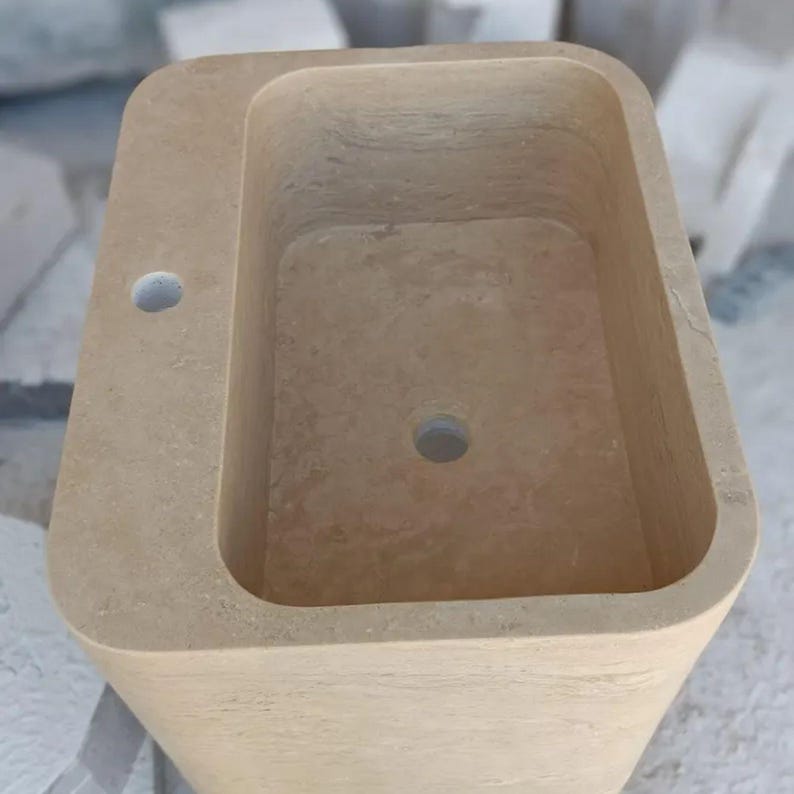 May include: A rectangular beige stone pedestal sink with rounded corners, featuring a smooth basin, a central drain hole, and a single faucet hole positioned on the flat deck area. The natural stone surface displays subtle variations in color and texture.