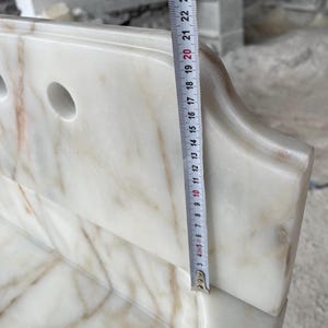 This luxurious Calacatta Gold marble sink features a standard drain hole with a back panel and a striking, elegant design with prominent grey and yellow veins on the front. It can be wall-mounted or countertop-mounted. The front has a fluted design. Dimensions: 24 inches long, 5 inches high, 14 inches deep, panel height 8 inches.