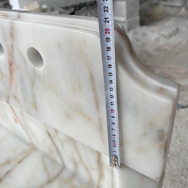 This luxurious Calacatta Gold marble sink features a standard drain hole with a back panel and a striking, elegant design with prominent grey and yellow veins on the front. It can be wall-mounted or countertop-mounted. The front has a fluted design. Dimensions: 24 inches long, 5 inches high, 14 inches deep, panel height 8 inches.