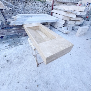 Handmade rectangular beige travertine stone sink with a recessed basin, thick solid edges, and natural stone texture, photographed in a marble workshop before bathroom installation.