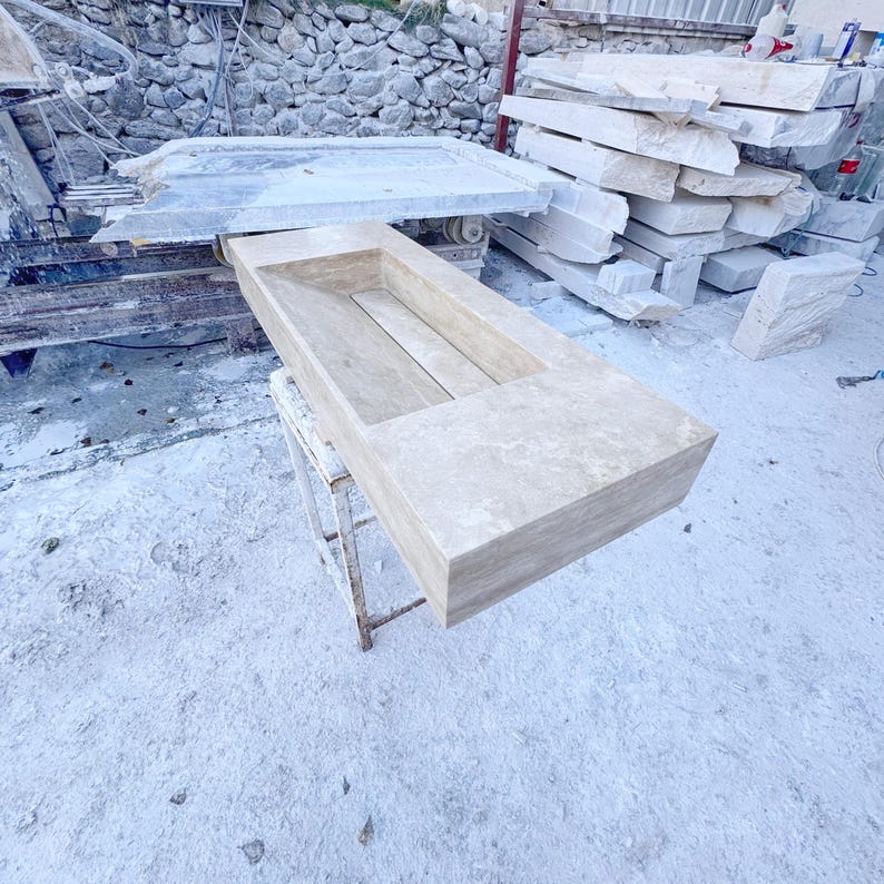 Handmade rectangular beige travertine stone sink with a recessed basin, thick solid edges, and natural stone texture, photographed in a marble workshop before bathroom installation.