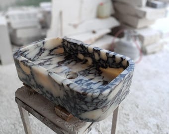 May include: A rectangular stone sink featuring a polished surface with a distinct marbled pattern of cream, white, and dark charcoal gray veins. The basin has rounded corners and a central circular drain hole, showcasing natural stone textures.