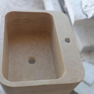 May include: A rectangular beige stone sink basin with rounded corners, featuring a smooth interior surface, a central circular drain hole at the bottom, and a smaller circular faucet hole positioned on the flat deck area to the right side.