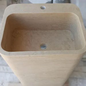 May include: A rectangular beige stone pedestal sink with a smooth, natural finish. The basin features a deep, hollowed-out center with a circular drain hole at the bottom and a single pre-drilled faucet hole centered on the flat top ledge surface.