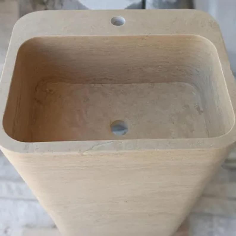 May include: A rectangular beige stone pedestal sink with a smooth, natural finish. The basin features a deep, hollowed-out center with a circular drain hole at the bottom and a single pre-drilled faucet hole centered on the flat top ledge surface.