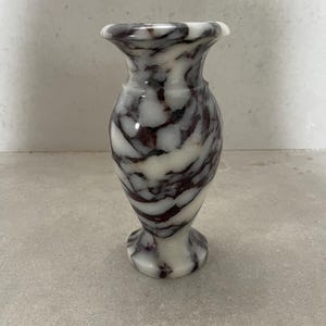 May include: A small, polished stone vase featuring a distinct marbled pattern with swirling veins of deep dark purple and white. The vase has a flared rim, a rounded body, and a circular pedestal base, showcasing a smooth, glossy finish throughout.
