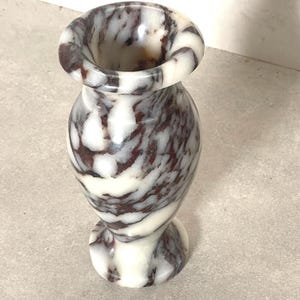 May include: A small, polished stone vase featuring a swirling, marbled pattern of deep burgundy and creamy white. The vase has a rounded, bulbous body, a narrow neck, and a flared, circular rim, with a matching circular base for stability.