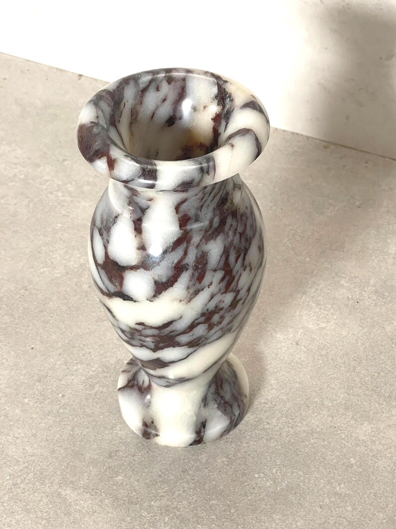 May include: A small, polished stone vase featuring a swirling, marbled pattern of deep burgundy and creamy white. The vase has a rounded, bulbous body, a narrow neck, and a flared, circular rim, with a matching circular base for stability.