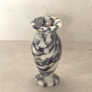 May include: A small, polished stone vase featuring a distinct marbled pattern with swirling veins of deep purple and creamy white. The vessel has a rounded body, a narrow neck, and a flared rim, standing on a circular base with a smooth finish.