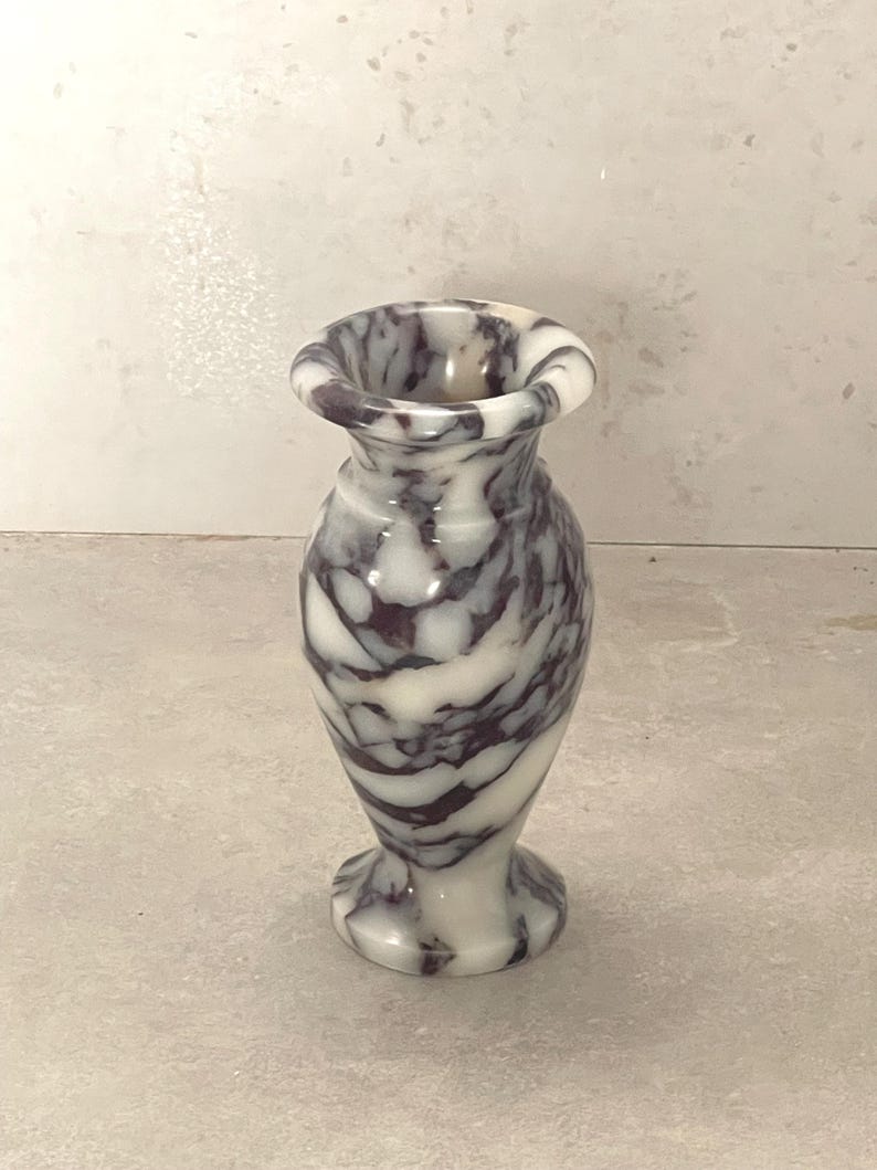 May include: A small, polished stone vase featuring a distinct marbled pattern with swirling veins of deep purple and creamy white. The vessel has a rounded body, a narrow neck, and a flared rim, standing on a circular base with a smooth finish.