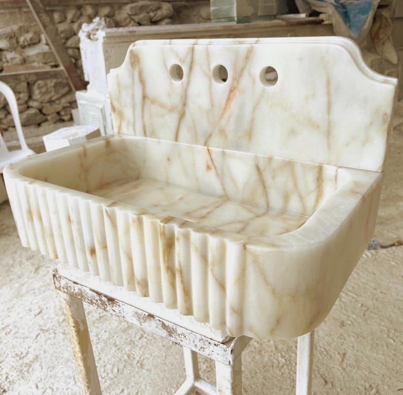 This luxurious Calacatta Gold marble sink with a back panel features a standard drain hole and a striking, elegant design with prominent grey and yellow veins on the front. It can be wall-mounted or countertop-mounted. The front has a fluted design.