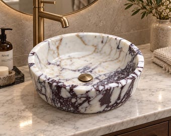 May include: A round vessel sink crafted from white marble with prominent deep purple and brown veining. The basin features a smooth, polished interior and exterior surface, complemented by a centered circular drain cover in a brushed brass finish.