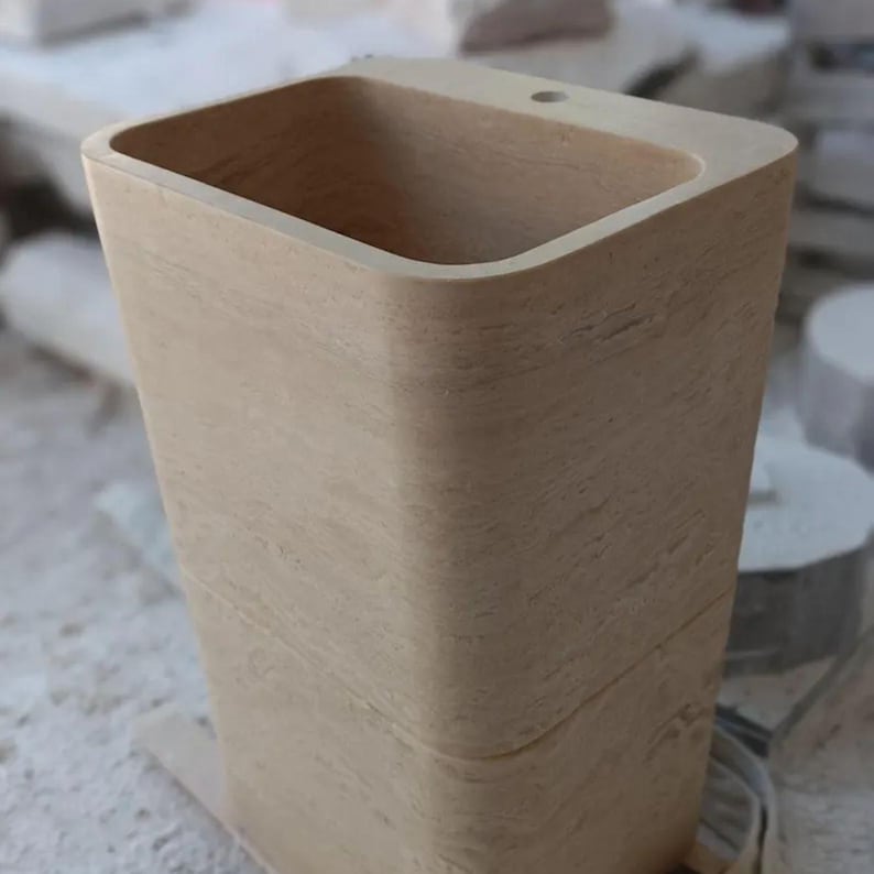 May include: A tall, rectangular beige stone basin with rounded corners and a smooth, textured surface. The basin features a deep, hollow interior and a single circular hole positioned on the top rim, showcasing natural stone grain patterns throughout.