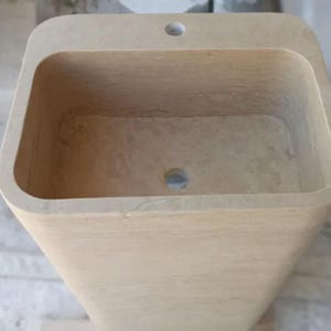 May include: A rectangular beige stone pedestal sink with a smooth, matte finish. The basin features a deep, hollowed-out center with a circular drain hole at the bottom and a single faucet hole centered on the flat top ledge of the basin rim.